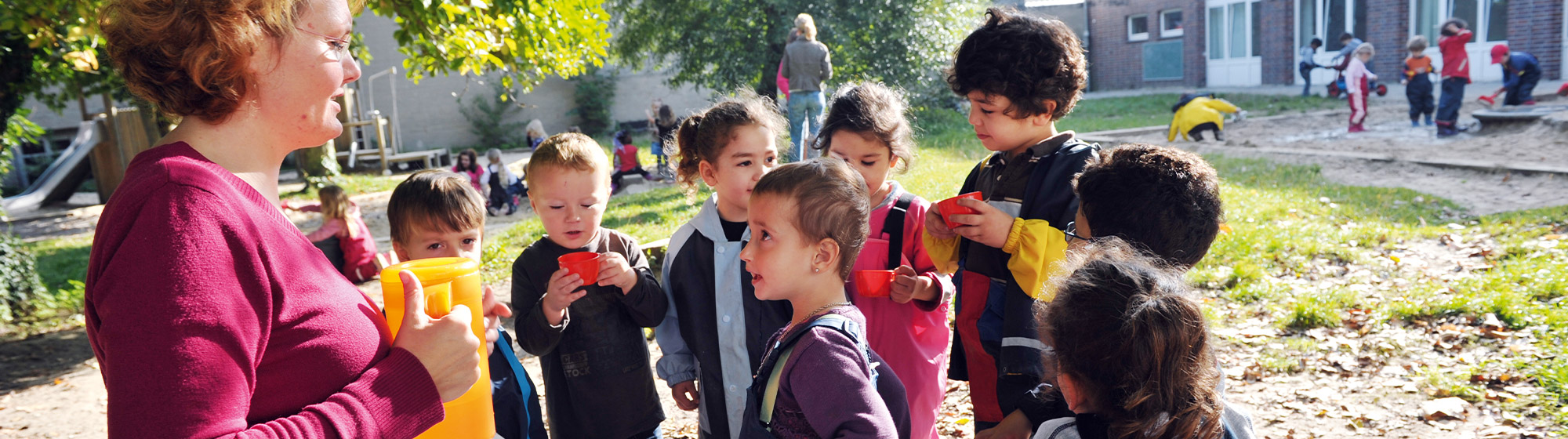 Kindergarten-Außengelände: Es ist gutes Wetter, die Sonne scheint. Im Vordergrund ist eine Erzieherin im Profil zu sehen, die eine Karaffe in der Hand hält. Um sie herum haben sich Kinder mit ihren Getränkebechern versammelt. Im Hintergrund sind weitere spielende Kinder und Erzieher:innen zu sehen.