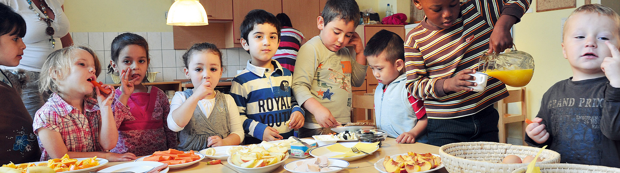 Kindergartenkinder stehen hinter einem Tisch, der mit gesunden Lebensmitteln wie Obst und Gemüse, Käse und Wurst eingedeckt ist. Ein Kind schenkt sich aus einer Karaffe Orangensaft in ein Glas ein. Einige der Kinder schlecken sich genüsslich die Finger ab. Im Hintergrund ist die Küche des Kindergartens zu sehen.