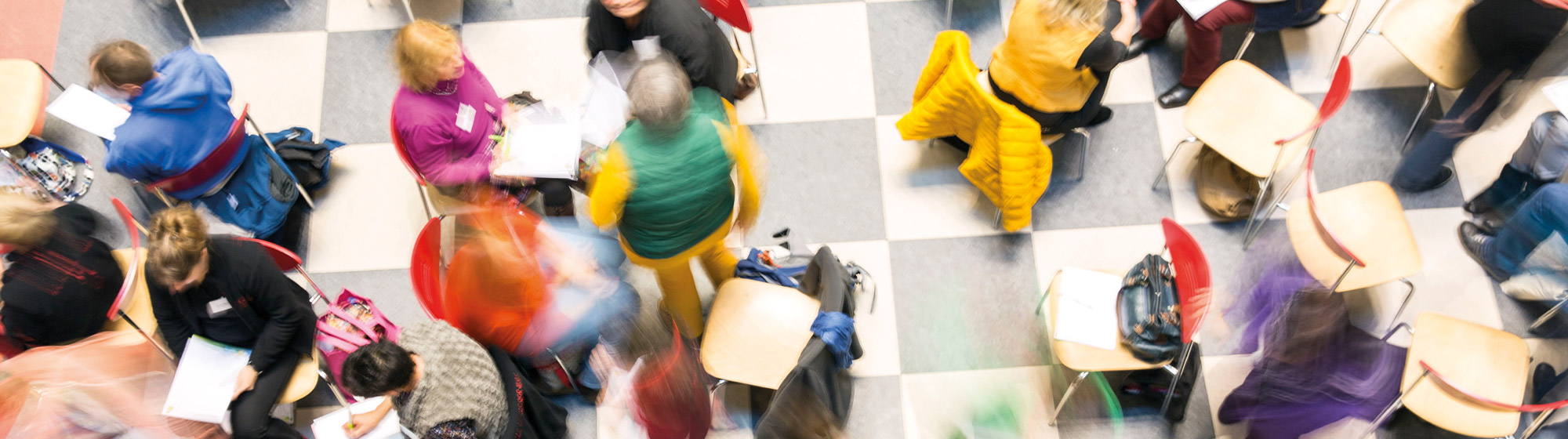 Das Bild ist auf einer ABmPE-Netzwerktagung entstanden. Es zeigt Menschen, die in Gruppen, auf bunten Stühlen, sitzen. Das Bild ist von oben herab fotografiert. Bewegungsunschärfe verzerrt das Bild optisch.